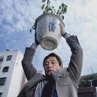 Mann hebt einen Blumentopf über den Kopf, Wasser tropft heraus. Im Hintergrund sind Wohngebäude und Himmel zu sehen.