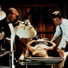 Zwei Männer in Anzügen stehen über einem Tisch mit einer Person unter einer Lampe. Ein großer Ziffernblock ist an der Wand.