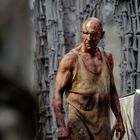 Kein Text erkennbar. Ein erschöpfter, von Schlamm bedeckter Mann in einem schmutzigen Unterhemd steht vor einer Texturwand.