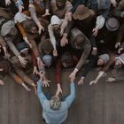 Viele Menschen in historischer Kleidung strecken ihre Arme einer Person auf Holzboden entgegen. Draufsicht.