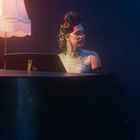 Keine erkennbaren Texte. Eine Person in elegantem Kleid an einem Flügel, mit retro Lampe daneben.