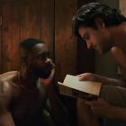 Zwei Männer, einer liest ein Buch vor, beide sind oberkörperfrei, in einem gemütlichen Raum mit Holzschrank.