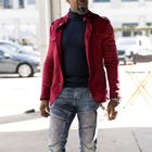 Keine sichtbaren Texte. Ein Mann trägt eine rote Jacke, dunklen Rollkragenpullover und Jeans. Hintergrund ist urban.