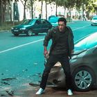 Eine verletzte Person steht neben einem beschädigten Auto auf einer leeren Straße. Autos im Hintergrund.