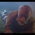 Ein blutverschmierter Mann kriecht in einer düsteren Umgebung über Gras, im Hintergrund eine verschwommene Lichtquelle.