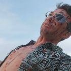 Ein Mann mit Sonnenbrille, blutverschmiertem Gesicht und offenem Hemd steht vor einem blauen Himmel.