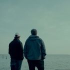 Zwei Männer stehen am Ufer und schauen aufs Wasser. Einer trägt eine Kappe, der andere eine Jacke. Ein Segelboot ist zu sehen.