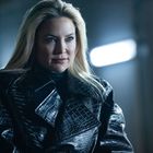 Keine sichtbaren Texte. Eine Frau mit blonden Haaren trägt einen glänzenden, dunklen Mantel. Die Umgebung ist dunkel beleuchtet.