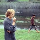 Ein Junge geht barfuß am Fluss entlang, während eine Frau in Gedanken versunken steht. Sie trägt einen groben Pullover.