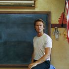 Ein Mann in weißem T-Shirt sitzt vor einer leeren Tafel in einem Raum mit US-Flagge.