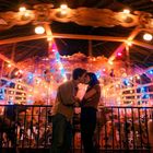 Ein Paar küsst sich vor einem hell erleuchteten Karussell mit bunten Lichtern, umgeben von einer nostalgischen Atmosphäre.
