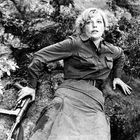 Keine sichtbaren Texte. Eine Frau liegt auf einem felsigen Untergrund, trägt eine militärische Kleidung und hält ein Gewehr.