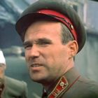 Zwei Männer in Militäruniformen, ein Mann mit rotem Band am Hut und einer mit Kopfverband, in einem zerstörten Hintergrund.