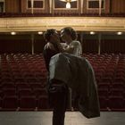 Ein eleganter Saal, großer Kronleuchter, leere rote Sitze. Paar steht auf Bühne, er trägt sie liebevoll.