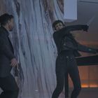 Zwei Personen in schwarzer Kleidung. Eine Frau schwingt ein Messer. Der Hintergrund zeigt eine marmorähnliche Wand.