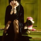 Eine Person mit dunklem Haar sitzt auf grünen Stufen, neben ihr ein Pudel mit rosa Details.