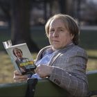 Ein Mann hält ein Buch mit dem Titel "Du bist kein Opfer" und sitzt auf einer Parkbank, blickt ernst in die Kamera.