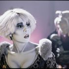 Eine Frau mit kurzen, blonden Haaren und blasser Haut schaut überrascht zur Seite. Hinter ihr ist eine verschwommene Figur.