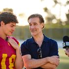 Ein junger Mann in Sportkleidung mit der Nummer 18, neben einem älteren Mann. Eine Frau hält ein Mikrofon in ihrer Hand.