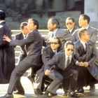 Es gibt keinen Text. Acht Männer in Anzügen, einige mit Sonnenbrillen, stehen vor einer Treppe in einer dramatischen Pose.