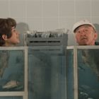 Keine Texte vorhanden. Eine junge Frau und ein älterer Mann blicken aus Aquarium-Tanks, umgeben von Fischen in einem kacheligen Raum.