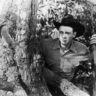 Ich kann nicht sagen, wer im Bild ist. Ein Mann mit Cowboyhut und Gewehr lehnt an einem Baum.