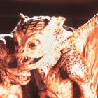 Zwei groteske Kreaturen mit schuppiger Haut, großen Augen und spitzen Zähnen. Leicht dämonisch, eng beieinander, in warmem Licht.