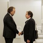 Zwei Männer im Anzug stehen in einem Waschraum und schütteln sich die Hände. Sie blicken sich ernst an.