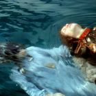 Ein Mensch treibt auf dem Rücken im Wasser, halb untergetaucht. Die Kleidung ist detailliert und verziert mit goldenen Scheiben.