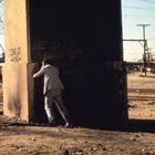 Graffiti auf Pfeiler: "CONEJO ANDA A..." und "VOTE NO ON BROWN". Drei Männer, einer lehnt an einem Betonpfeiler. Schrottplatzumgebung.