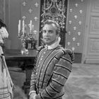 Ein Mann in historischer Kleidung steht in einem dekorativen Raum, ein Kerzenständer steht auf einem Tisch im Hintergrund.