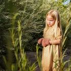 Ein Mädchen steht nachdenklich im Freien, umgeben von hohen Pflanzen. Sie trägt ein langes Kleid und gestrickte Handschuhe.