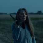 Eine Frau mit langem Haar hält eine Waffe auf der Schulter, vor einer weitläufigen, dämmerigen Landschaft.