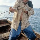 Ein Mann in zerknittertem Hemd hält ein Seil auf einem Holzbötchen im Wasser, sein Gesicht wirkt angestrengt.
