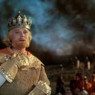 Ein Mann mit Krone und Rüstung blickt nachdenklich; im Hintergrund Soldaten und Rauch am Himmel.