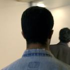 Zwei Personen gehen in einem Flur, eine im Vordergrund von hinten sichtbar.