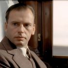 Es ist ein Mann in einem Anzug, der in einem Zugabteil sitzt und ernst schaut.