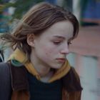 Es gibt keinen Text. Ein junges Mädchen mit braunen Haaren und Rucksack sieht nachdenklich aus und geht an einem weißen Zaun vorbei.