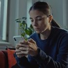 Eine Frau sitzt nachdenklich auf einem roten Sofa und schaut auf ihr Smartphone. Im Hintergrund ist ein Fenster.