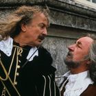 Zwei Männer mit langen Haaren und Schnauzbärten in historischer Kleidung, einer hebt ein Schwert, schauen sich intensiv an.