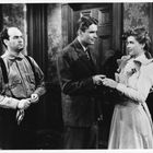 Drei Personen in einem Raum. Ein Mann links verschränkt die Hände, während ein Paar sich anblickt und die Hände hält.