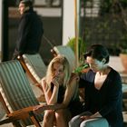 Zwei Frauen sitzen auf Liegestühlen, eine tröstet die andere. Ein Mann steht im Hintergrund. Es ist Abend.