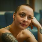 Eine Frau mit Glatze lächelt sanft, stützt ihren Kopf auf die Hand. Sichtbare Tattoos auf Schulter und Arm, gemütliche Umgebung.