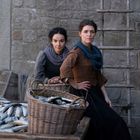 Zwei Frauen in historischer Kleidung neben Körben voller Fische, vor einer Steinmauer mit Leiter.