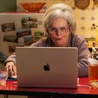 Eine ältere Frau sitzt konzentriert vor einem Laptop mit einem angebissenen Apfel-Logo. Dazu ein Glas und eine Flasche.