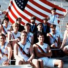 Sportler in weißen Outfits mit US-Flaggen-Emblem sitzen auf Tribünen. Eine große US-Flagge wird im Hintergrund gehalten.