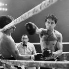 Zwei Boxer im Ring, einer schlägt zu, der andere weicht aus. Ein Schiedsrichter beobachtet aufmerksam. Boxhandschuhe sichtbar.