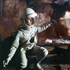 „DEFA“-Logo unten rechts. Ein Astronaut in einem Raumanzug kniet in einer futuristischen, metallischen Umgebung.