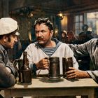 Ein Seemann in weißem Hemd hält einen Krug, begleitet von zwei weiteren Männern in maritimer Kleidung in einer Taverne.