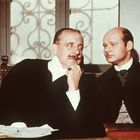 Zwei Männer in formeller Kleidung sitzen nebeneinander vor einem Fenstergitter. Der linke Mann gestikuliert nachdenklich.